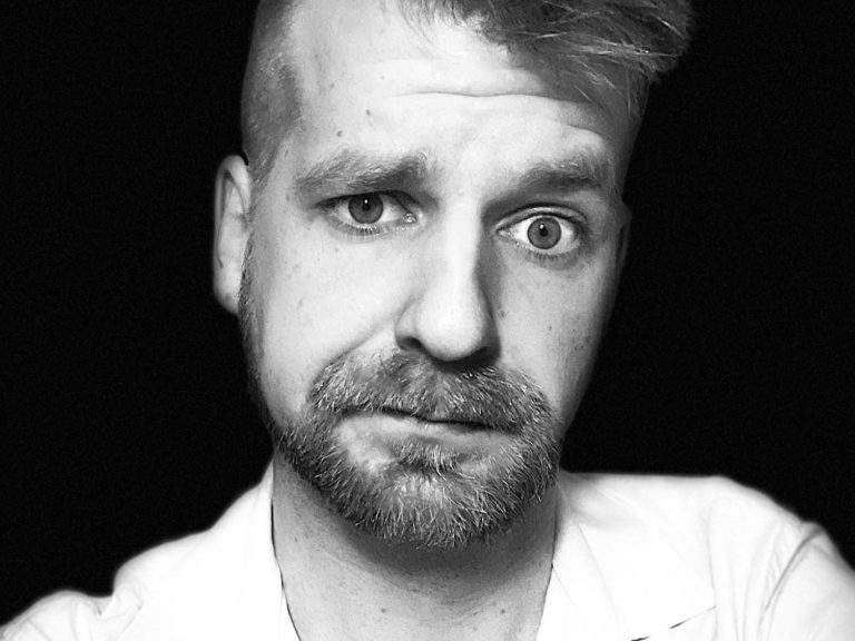 Ein Portrait von Arty Schulz im weißen Hemd, mit Vollbart und freundlichem Blick, Haare zur Seite gestylt.
